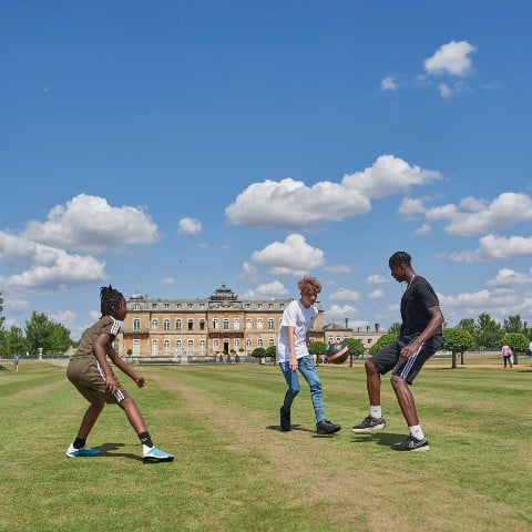 Three young men are kicking a football on grass in front of Wrest Park, a mansion in the style of a French chateau