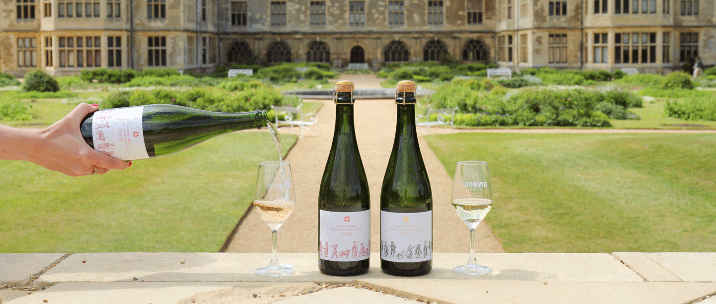 Two bottles of wine are stood on a stone ledge next to two full glasses of wine. They are positioned in front of a large stately home. Someone's hand is in view, pouring a glass of wine from one of the bottles.