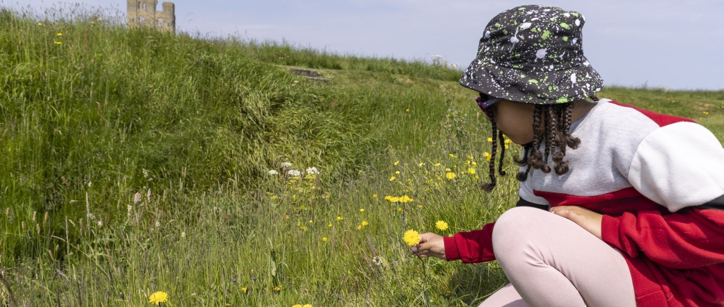 Photo of a child wearing a hat and sunglasses looking at a dandelion with Scarborough Castle in the distance