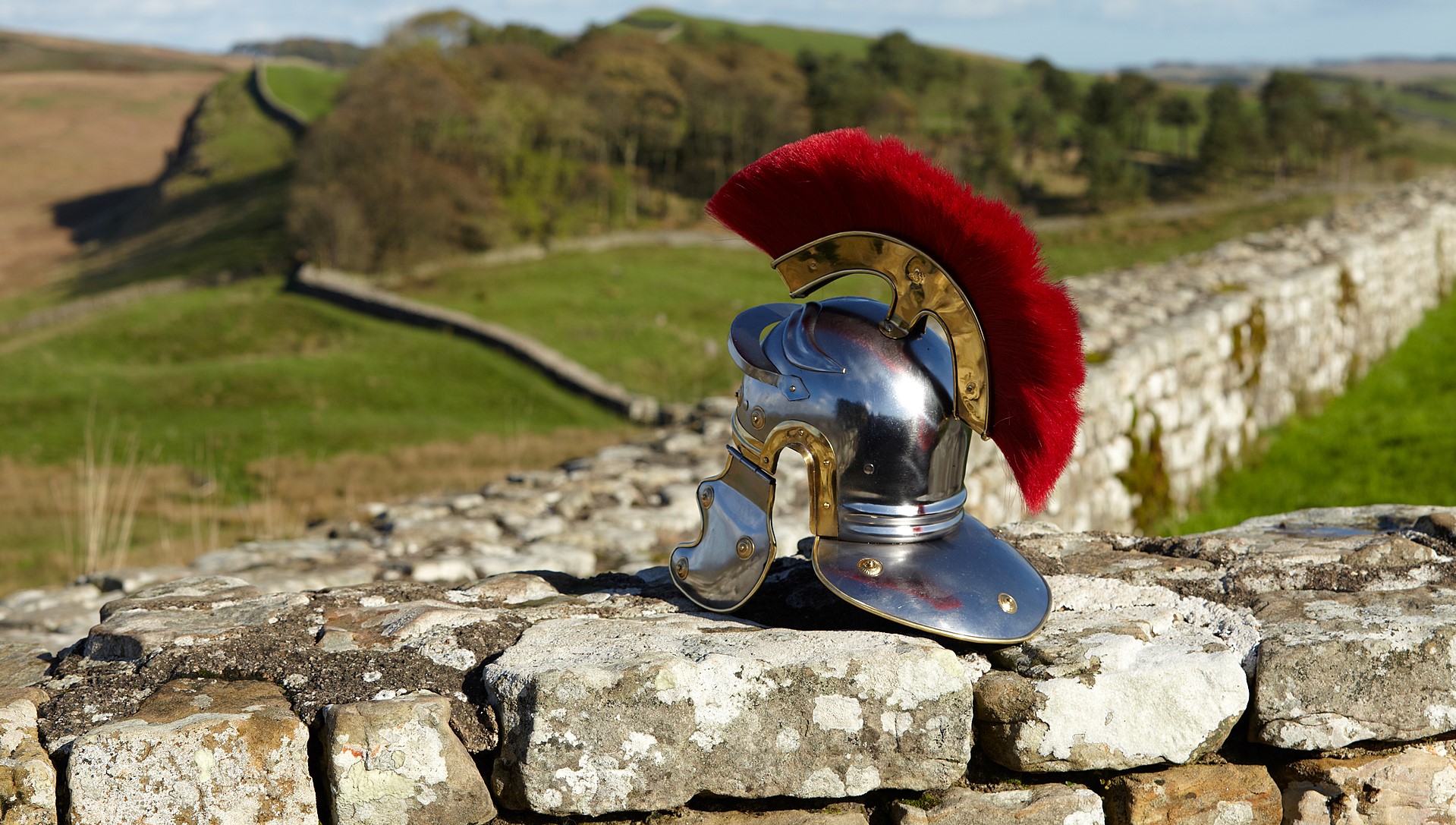 A metal Roman helmet with a dark red feather coming out of the top rests on a section of stone wall.