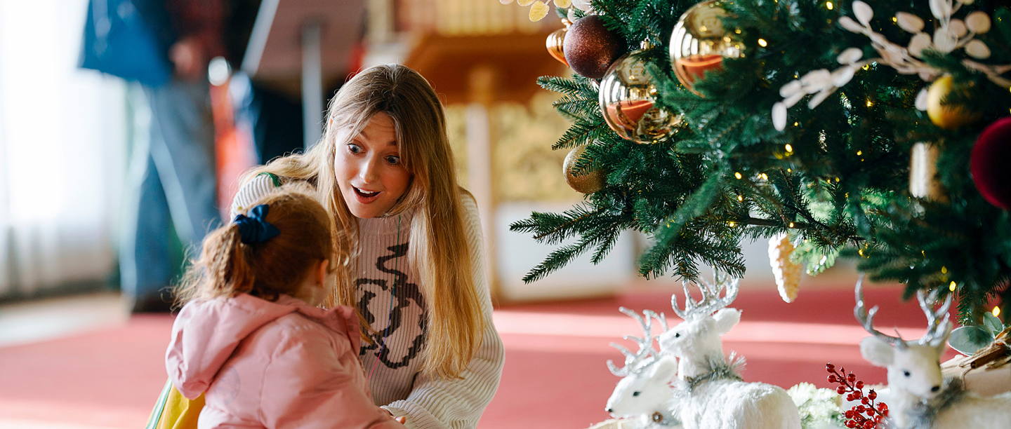 An adult and a young child sit on a red carpet beside a decorated Christmas tree, looking at festive decorations and white reindeer figurines at Wrest Park. 