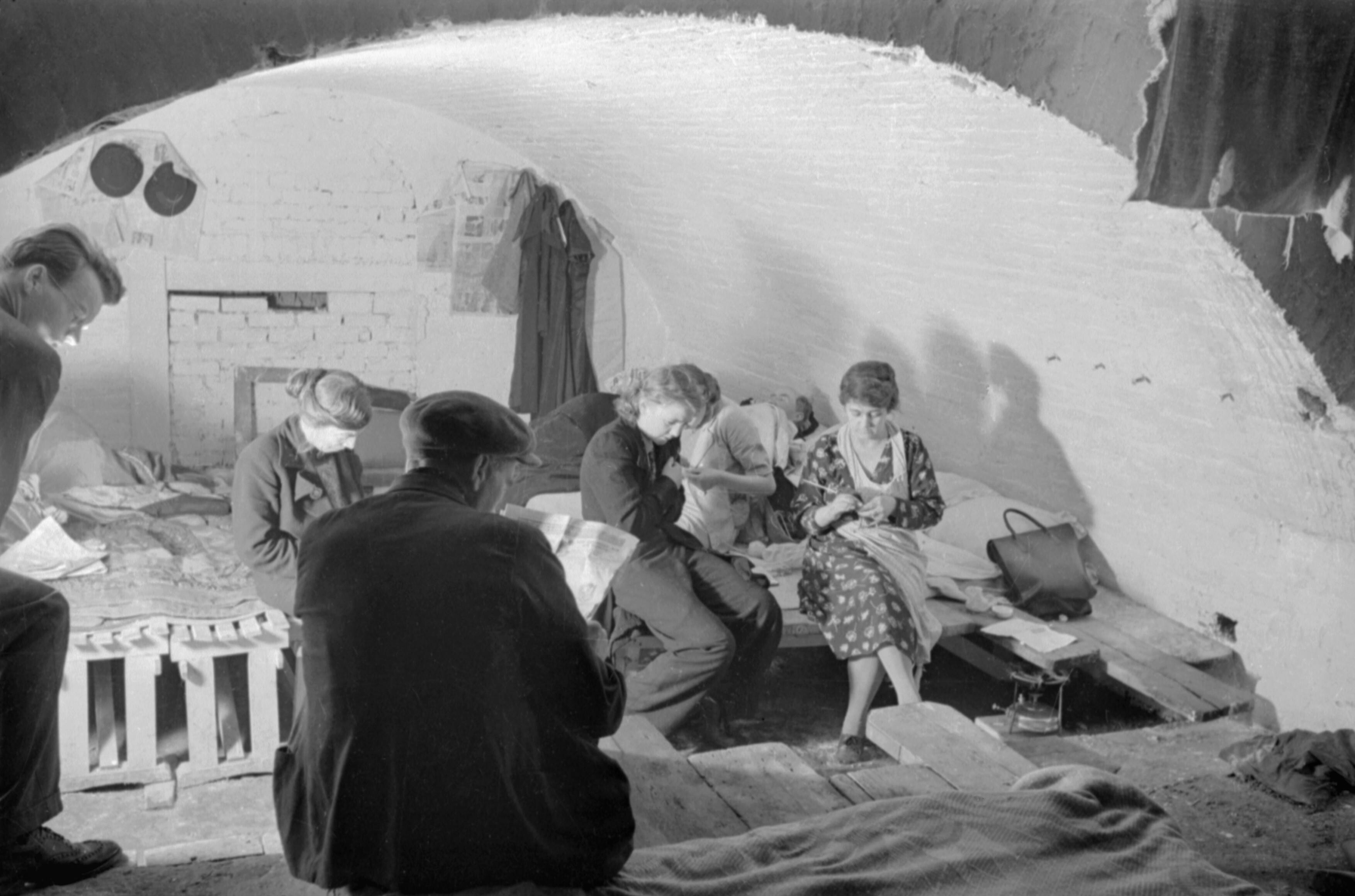 A small group of shelterers knit and read the evening newspaper in this small section of an air raid shelter under the railway arches, somewhere in South East London. Makeshift beds have been constructed from crates and planks of wood. This photograph was probably taken in November 1940.