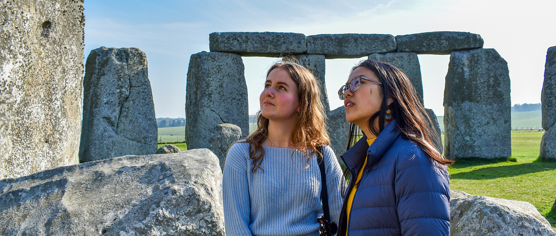 Two young women look up close at the stone circle at Stonehenge on a sunny day.