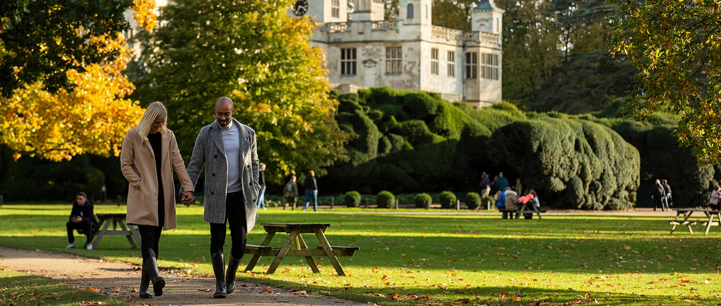 Two adults wearing long coats walk through the gardens at Audley End smiling with autumn leaves on the trees in the background.