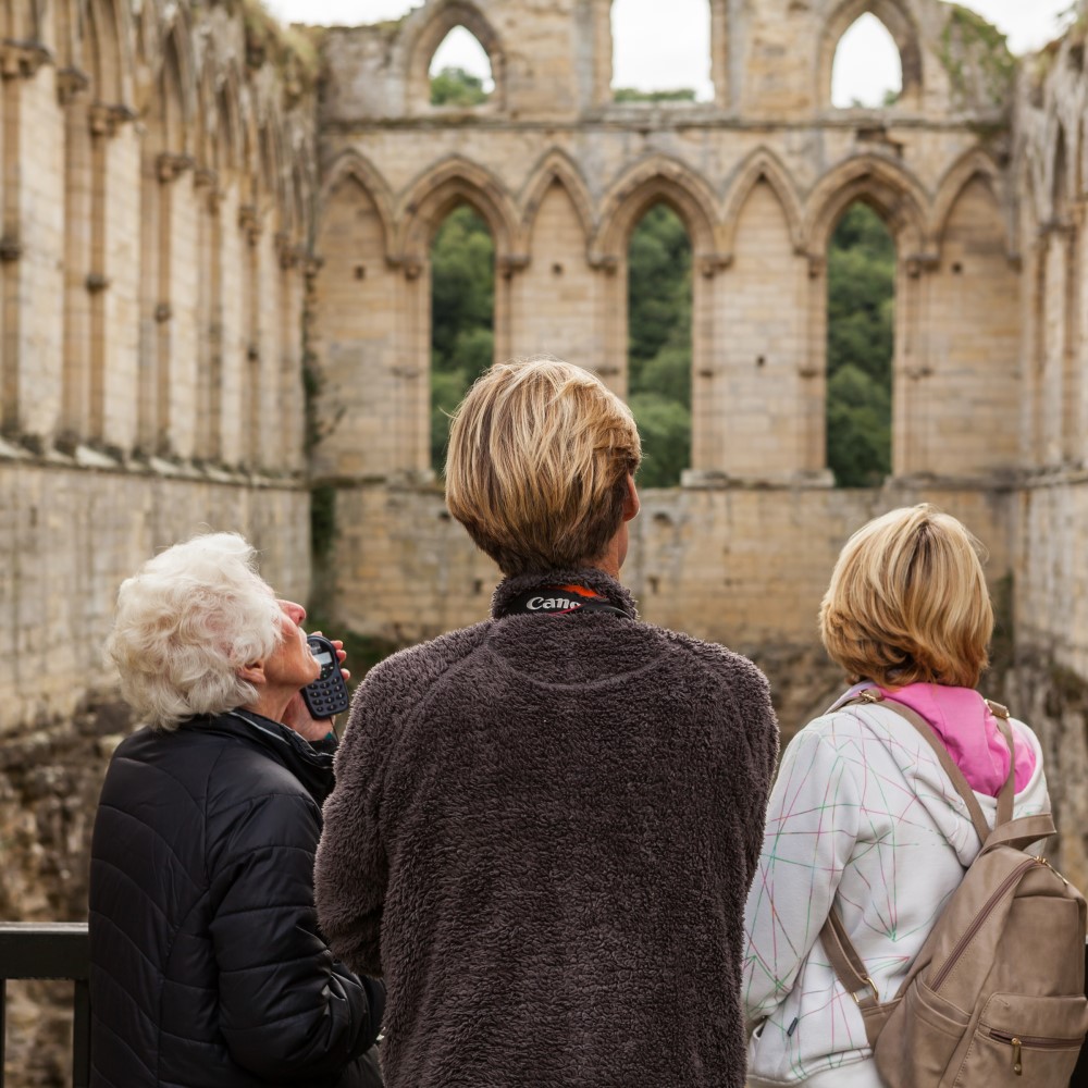 Photo of three people looking out at the ruins of Rievaulx Abbey on a cloudy day