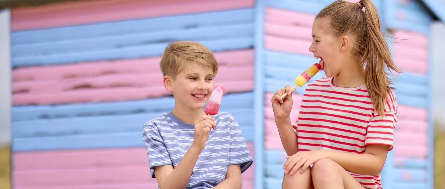Photo of two children eating ice lollies in front of a beach hut