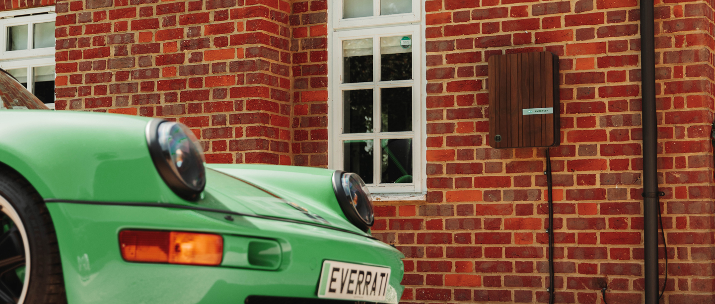 A green car next to an Andersen EV charger which is mounted to a red brick wall.