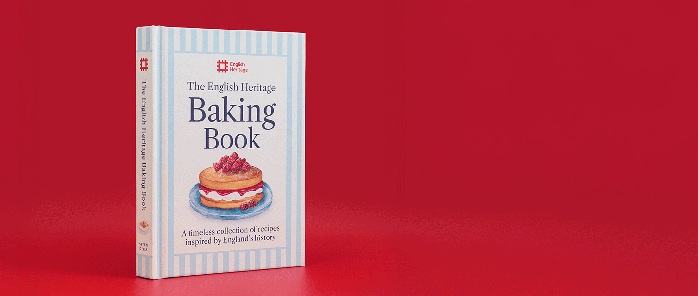 A hardback cream and blue book stood upright on a plain red background. The book is titled ' The English Heritage Baking Book and features a colourful illustration of a victoria sponge cake.