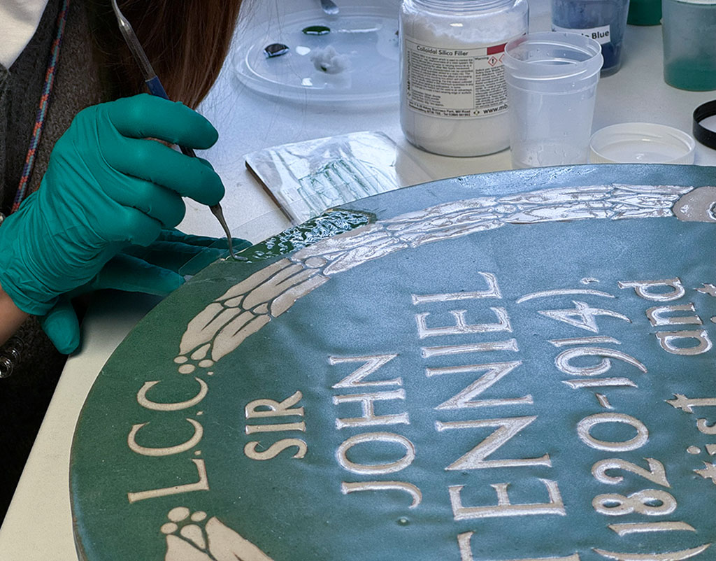 Colour photograph showing conservator's hands filling a damaged part of a jade green plaque for John Tenniel