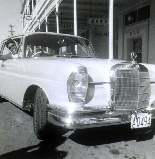 Black and white photograph of Winifred Atwell wearing a headscarf and smiling and waving from the driving seat of a light-colour Mercedes with shops and bars in the background. Black and white photograph of Winifred Atwell wearing a headscarf and smiling and waving from the driving seat of a light-colour Mercedes with shops and bars in the background.