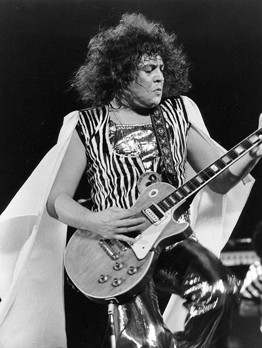 Black and white three quarter length portrait of Marc Bolan playing the guitar and dancing whilst wearing a floaty white cape, a zebra-print zip up waistcoat over a silver lamé jumpsuit