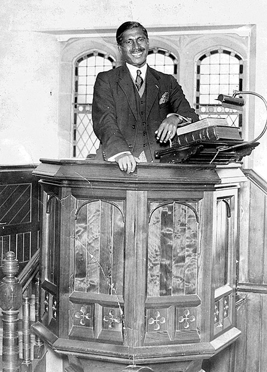 Black and white photograph of Kamal Chunchie in a carved wood pulpit, leaning on a lectern