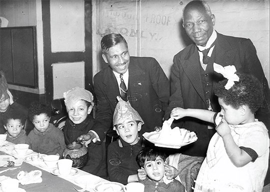Black and white photograph of pastor Kamal Chunchie pouring tea from a pot into the cup of a child at a table, while another adult serves them a platter of sandwiches. Some of the children pull faces and wear paper hats.