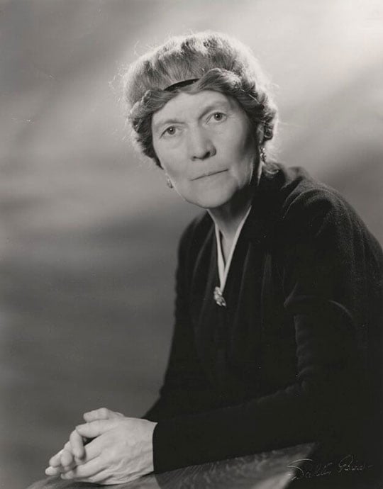 Black and white half-length photograph of Ivy Compton-Burnett, a white woman with a black dress, clasped hands, and a distinctive hairstyle with hair wound around a band around her head.