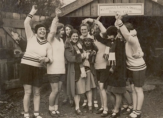 Black and white photograph of cheering women outside a boatshed