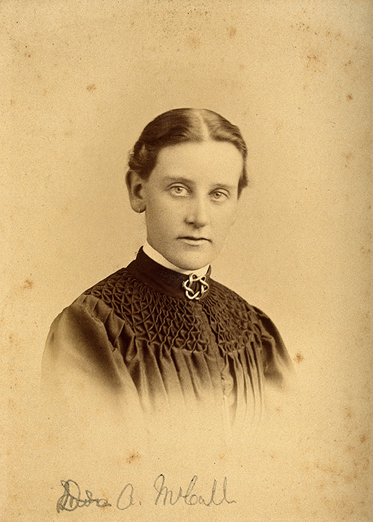 Sepia-toned portrait of Dr Annie McCall with a black smocked dress, brooch at her throat and parted, tied-back dark hair