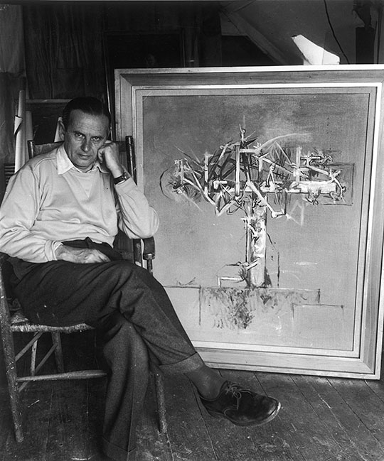 Black and white portrait of artist Graham Sutherland sitting in front of a crucifix painting