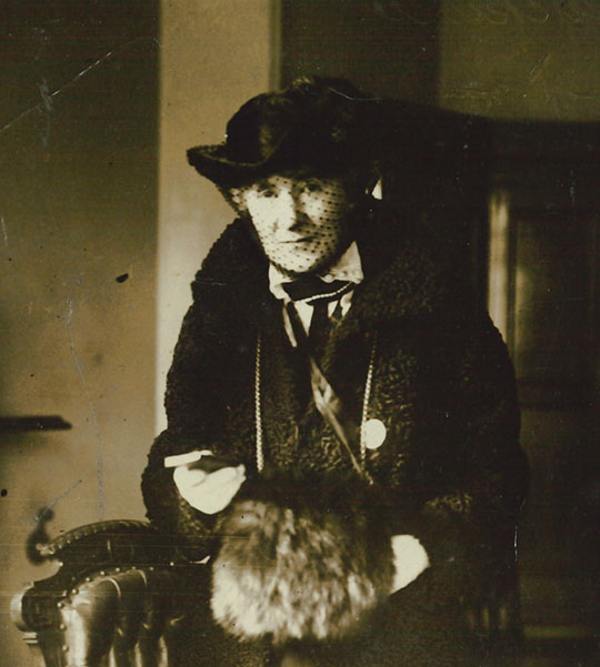Portrait of Gertrude Tuckwell with a black hat with mesh veil, fur stole and dark patterned coat. She holds a book in her right hand.