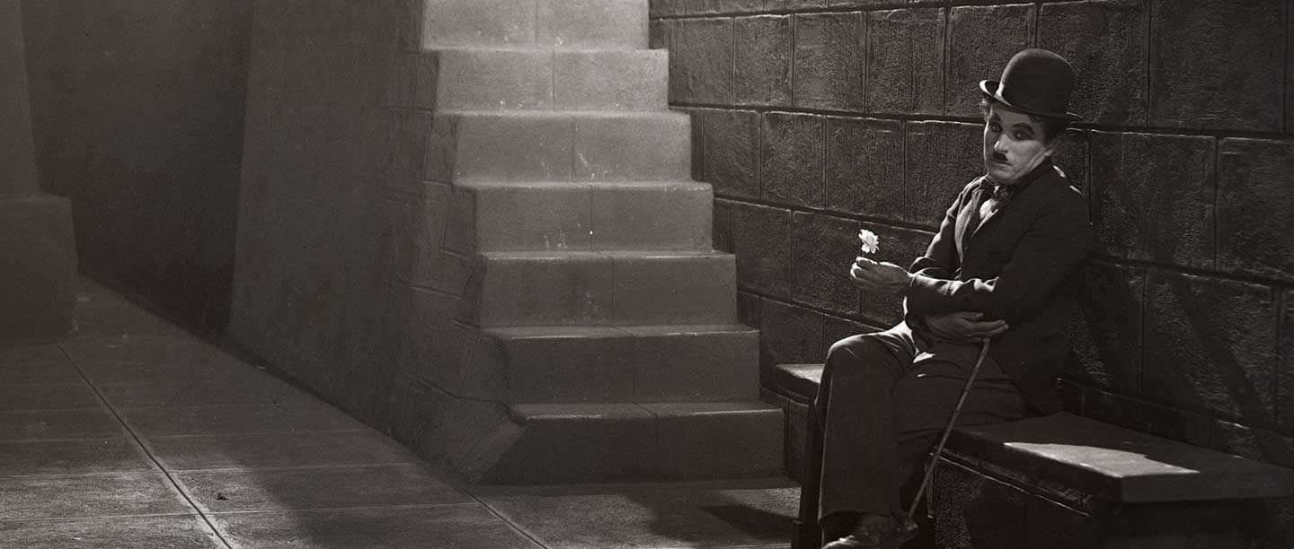 Black and white still of Charlie Chaplin sitting on bench holding flower
