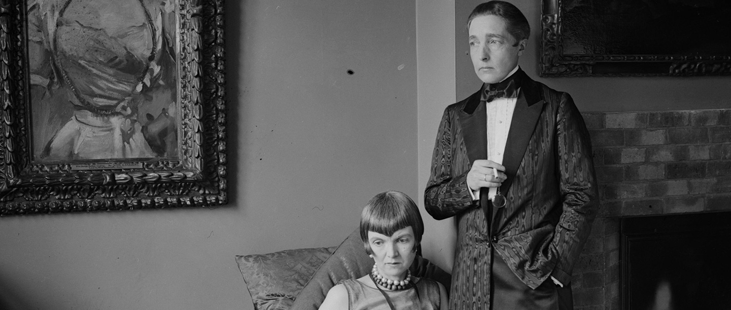 Black and white photograph of Radclyffe Hall in men's dinner jacket standing beside Lady Una Troubridge, seated and wearing evening dress with pearl necklace 