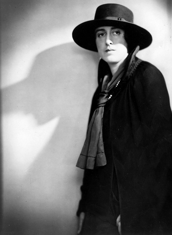 Black and white photograph of Vita Sackville-West standing at right of frame, wearing dark cloak and broad-rimmed hat 