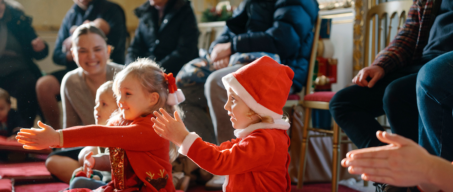 Two young children dressed up in Father Christmas outfits and hats smile and clap on the floor next to a group of adults at Wrest Park. 