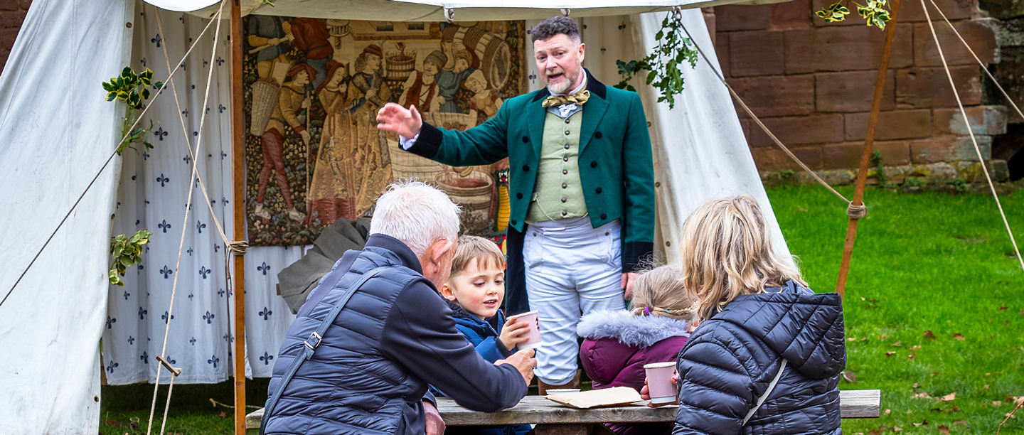 A man dressed in a dark green tailored jacket and green waistcoat performs in front of adults and children in a tent at Kenilworth Castle.