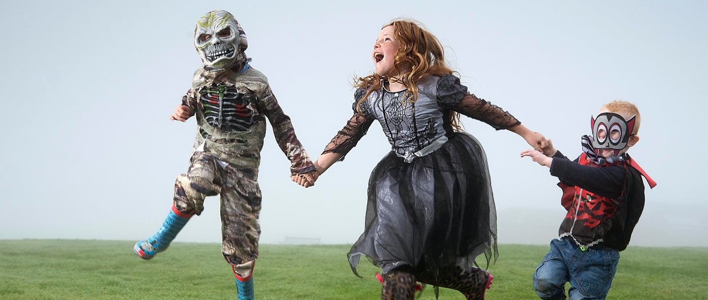 Three children jump in the air holding hands wearing Halloween costumes on a foggy day.