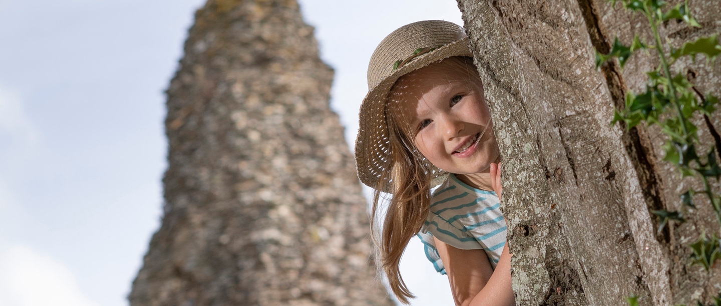 A young blonde girl in a sunhat peeps around the old walls of Launceston Castle. She is smiling.