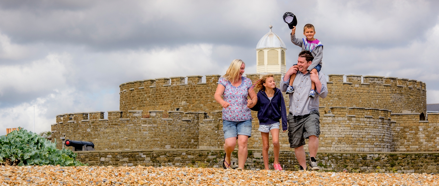 A mum and dad walk down the beach in front of Deal Castle in summer with their young son and daughter. The dad is carrying his son on his shoulders, and his son swings his cap in the air. 