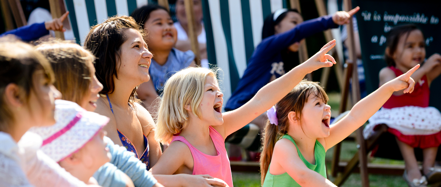 A group of children accompanied by adults sit on deckchairs pointing at a performer.