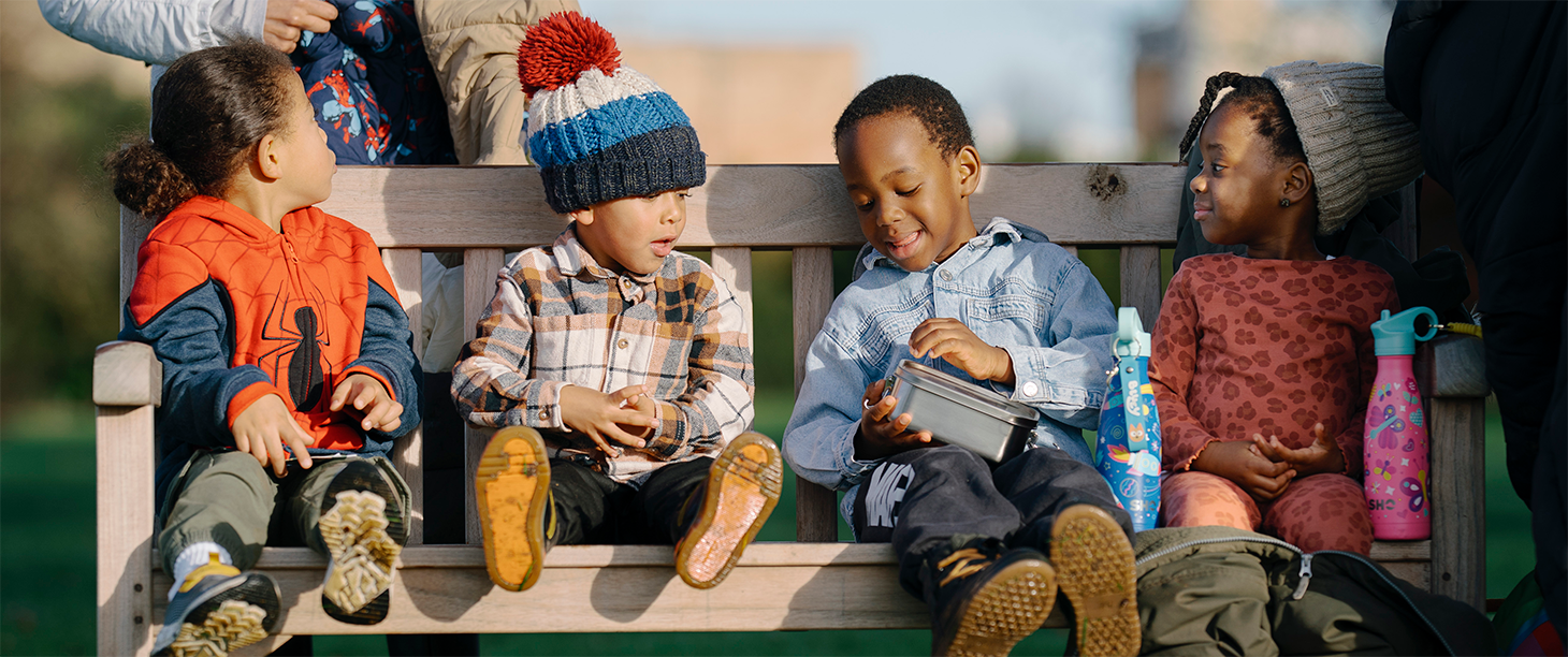 Image: four children sit on a bench in outdoor clothing, having snacks and drinks