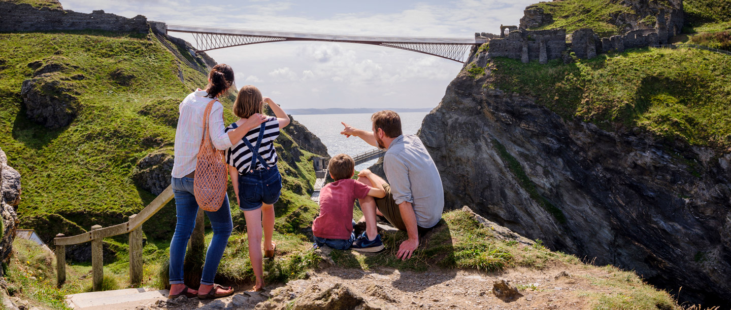 Two adults and children stand overlooking Tintagel Castle and bridge on a sunny day