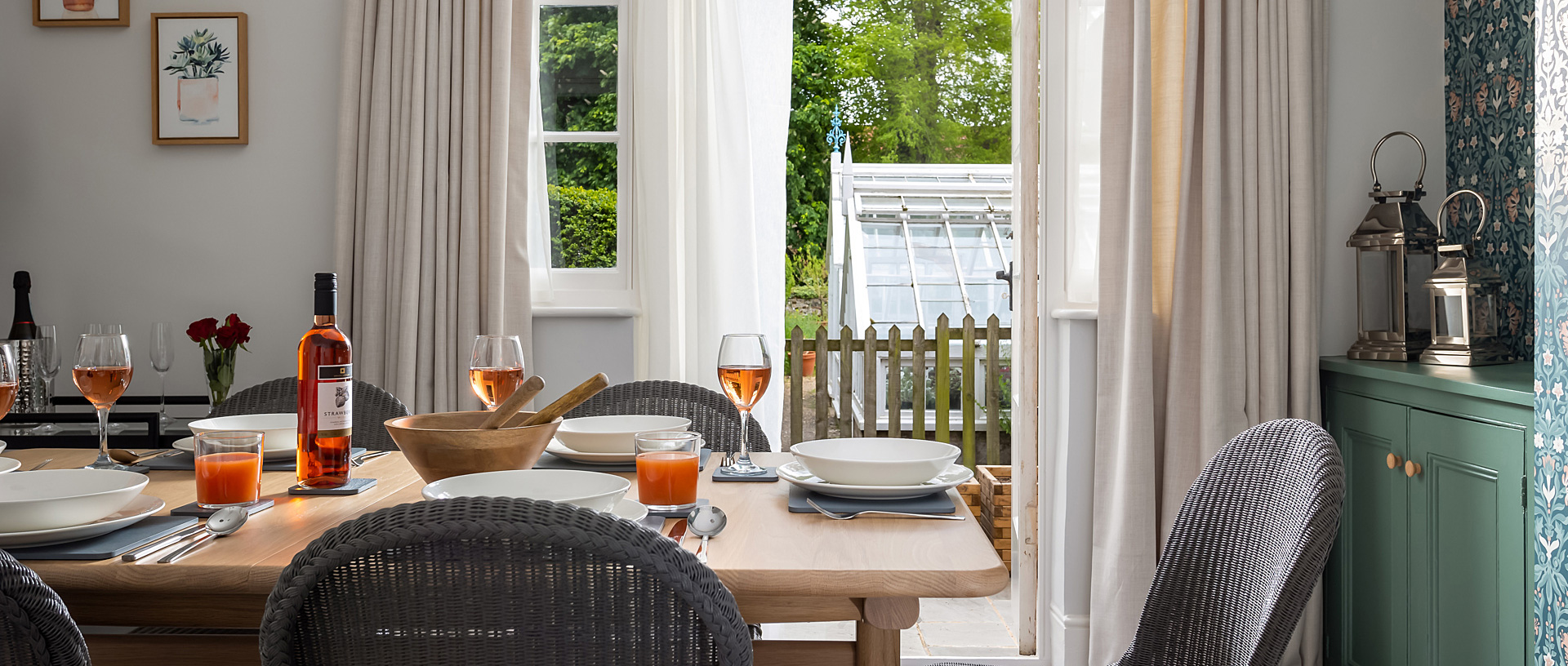 A wooden table laid out with plates and filled wine glasses, with view of the garden in a holiday cottage.