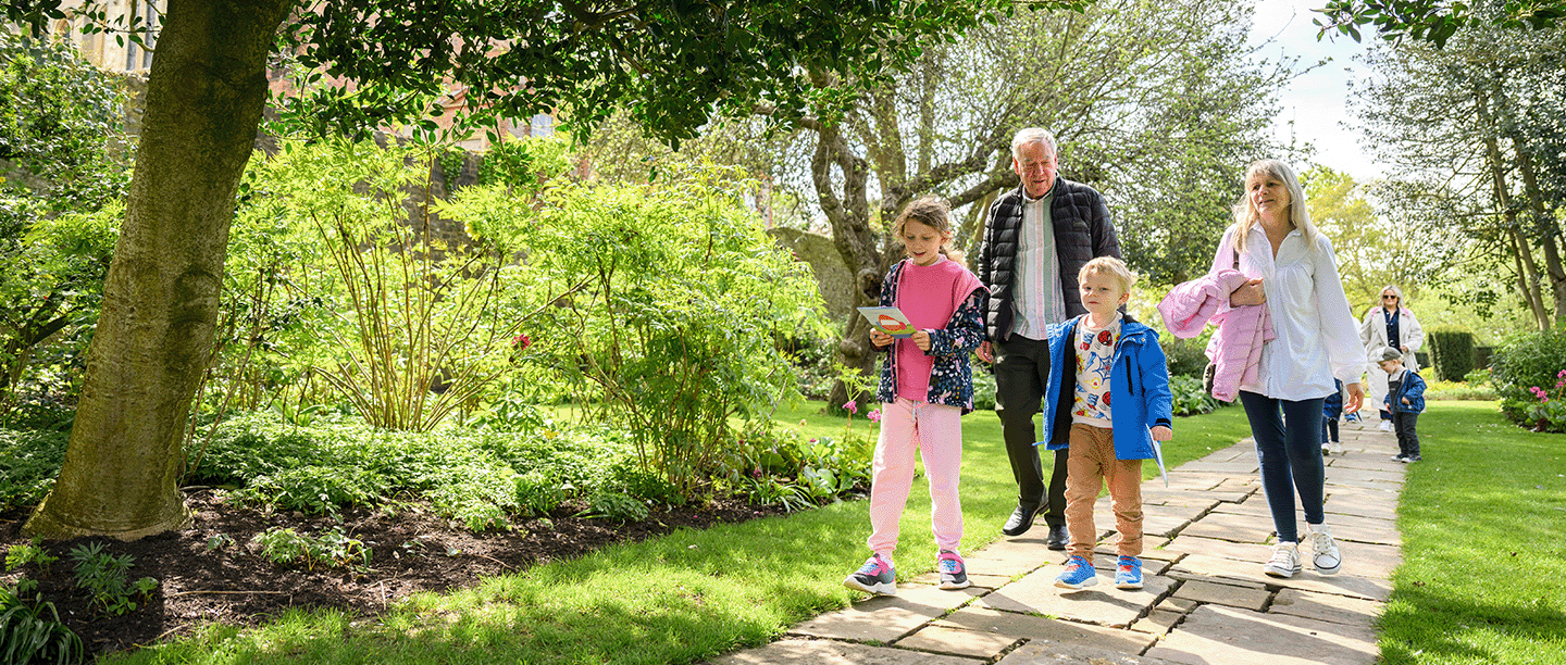 Photo of two adults and two children holding Easter adventure quest leaflets and walking down a path at Eltham Palace and Gardens in London