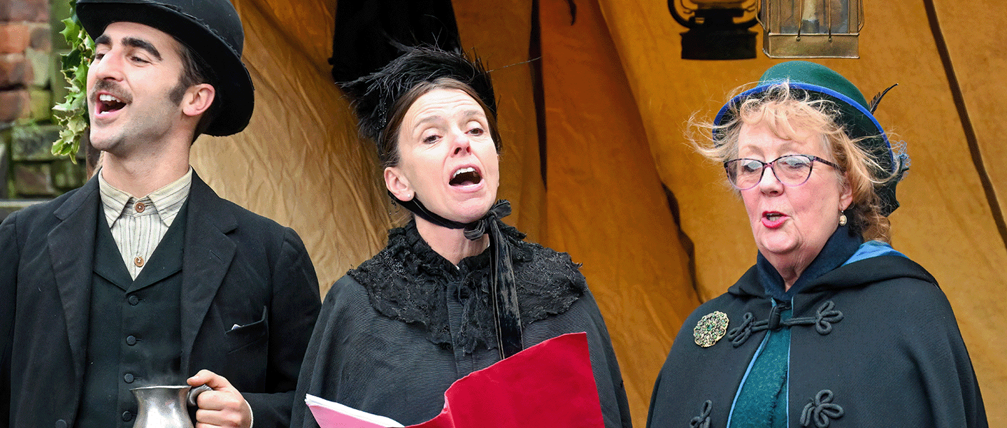 Photo of three people dressed in Victorian clothing and singing Christmas carols