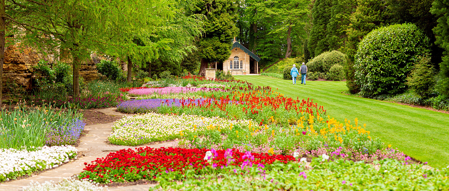 A view of two people in the distance walking towards a garden outbuilding, known as the Target House at Brodsworth Hall. There are a variety of colourful flowers growing in the flower beds. 