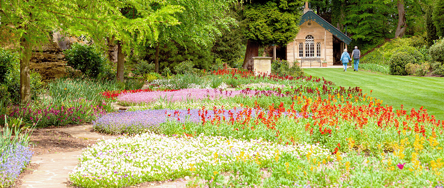 Photo of two people walking towards the Target House next to colourful parterre gardens and woodland at Brodsworth Hall and Gardens in South Yorkshire