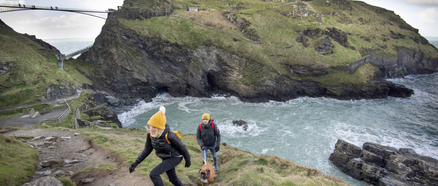 Photo of two people in winter clothing and a dog walking up a slope at Tintagel Castle in Cornwall