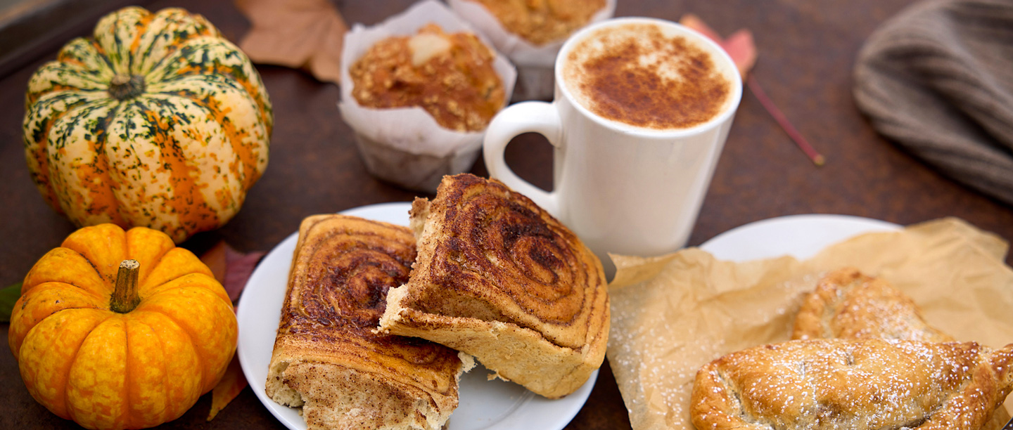 A close up of cinnamon buns, sweet pastries and muffins on a wooden table positioned next to two small pumpkins and a hot drink.