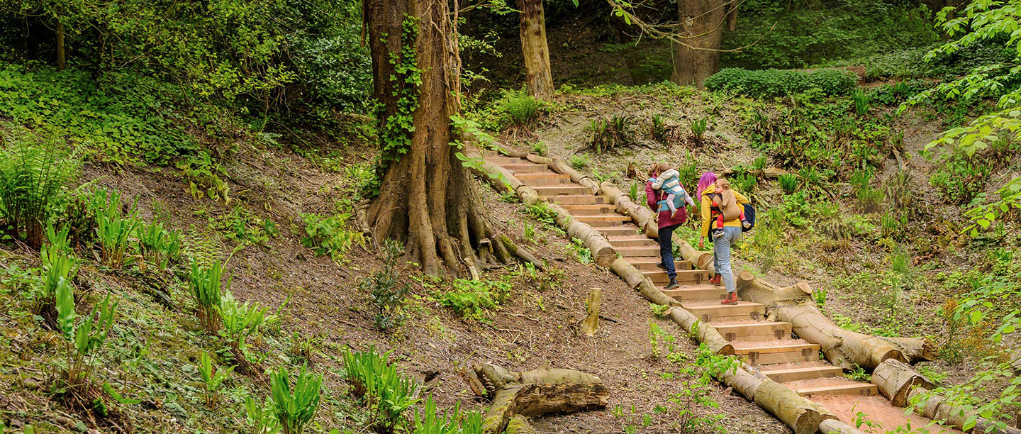 Photo of two adults, each carrying a toddler on their back, walking up a set of steps in a historic garden