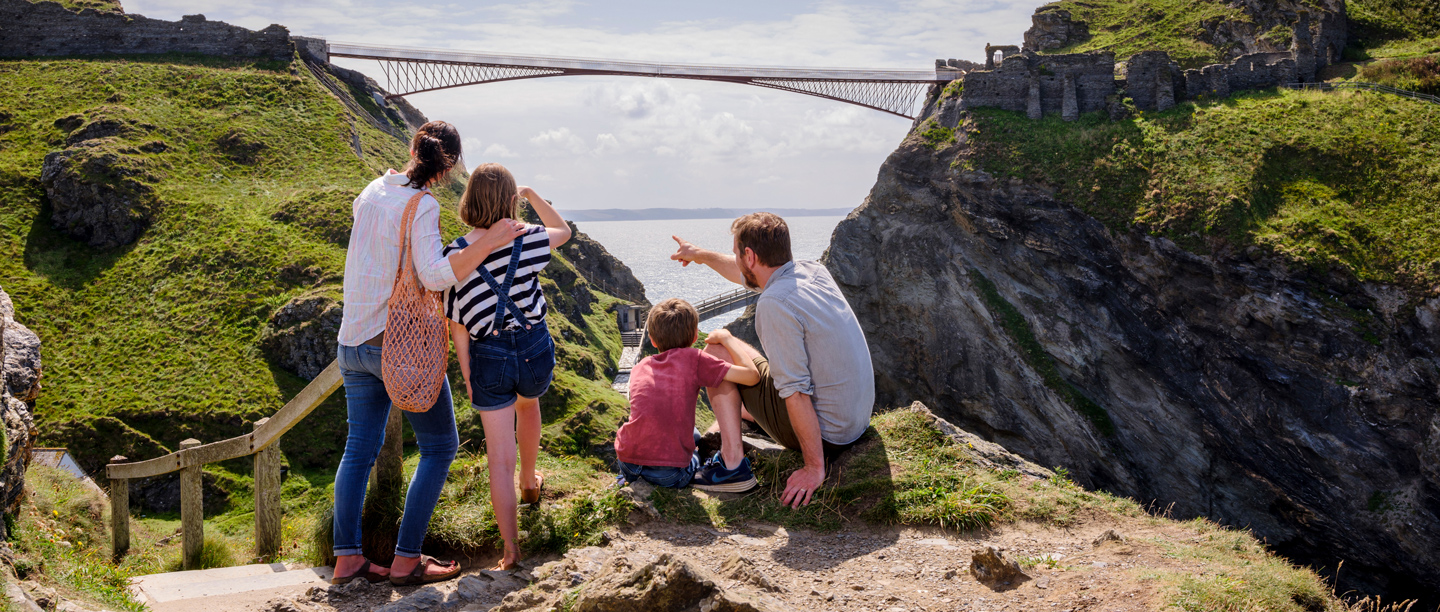 Two adults and children point and look towards the bridge at Tintagel Castle on a sunny day.