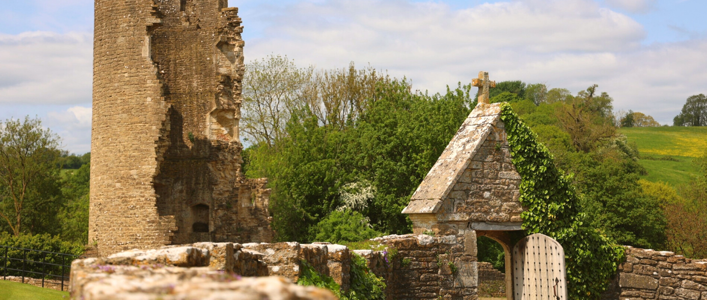 The stone ruins of Farleigh Hungerford Castle surrounded by green trees and grass under a partly cloudy sky.