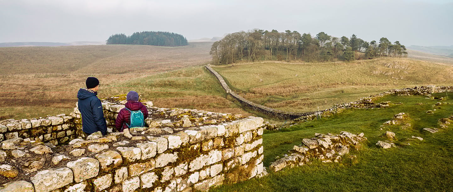 A man and a woman dressed in coats stand overlooking a section of Hadrian's Wall at Housesteads Roman Fort. 