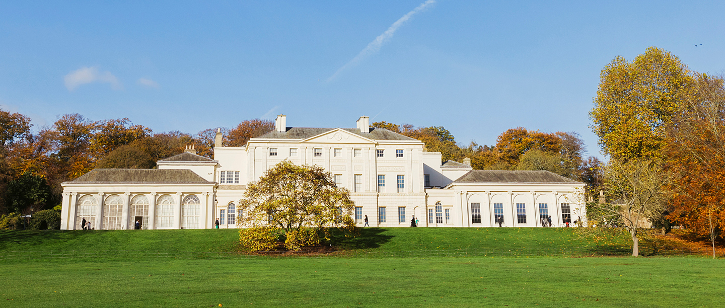 A view of Kenwood House on a sunny day surrounded by trees with orange and brown coloured leaves.