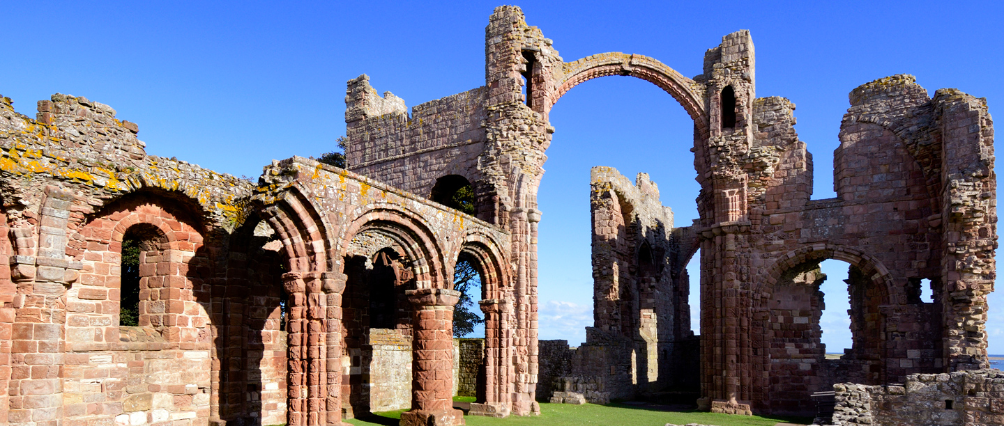 A view of the ruins of Lindisfarne Priory walls and arches under a blue sky.