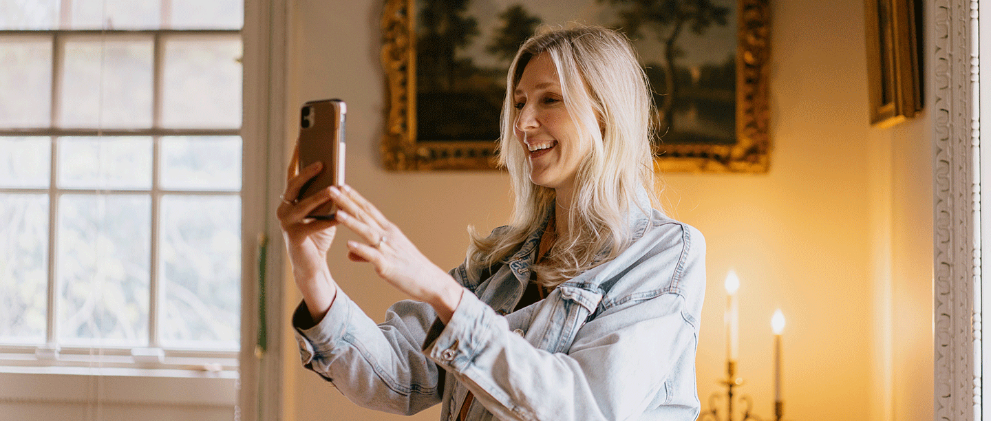 A woman holding a phone up to take a photograph. The woman is smiling and is stood inside a grand room at Marble Hill House.