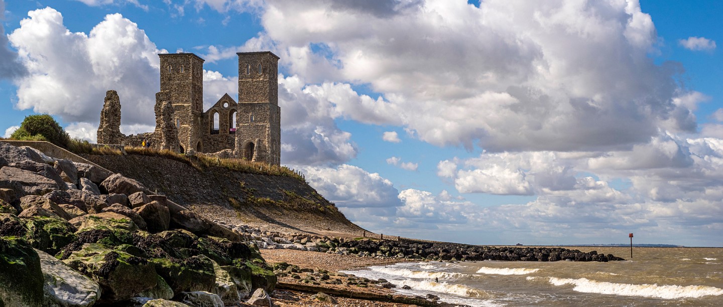 Ruins of a twin‑towered medieval church stand on a grassy headland above a rocky beach, overlooking choppy waves under a bright sky filled with large, billowing clouds.