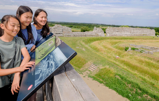 Photo of an adult and two children looking out over Richborough Roman Fort and Amphitheatre on a sunny day Photo of an adult and two children looking out over Richborough Roman Fort and Amphitheatre on a sunny day