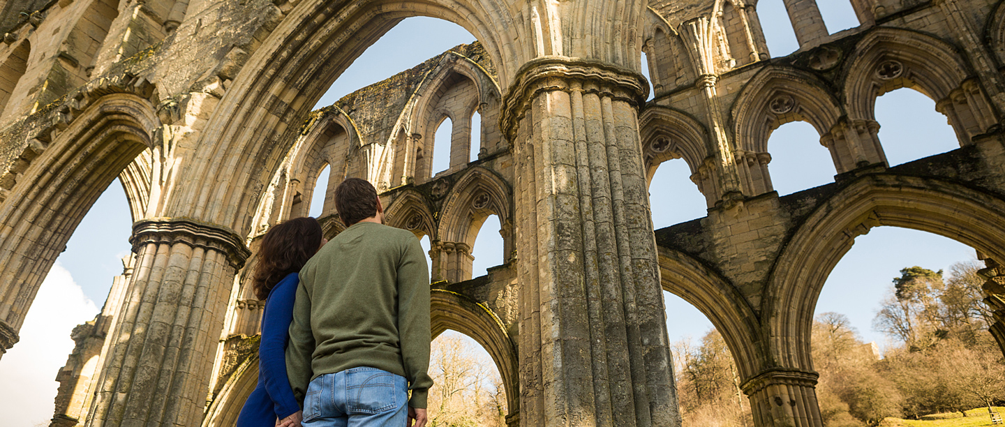 Two adults stand looking up at Rievaulx Abbey ruins.